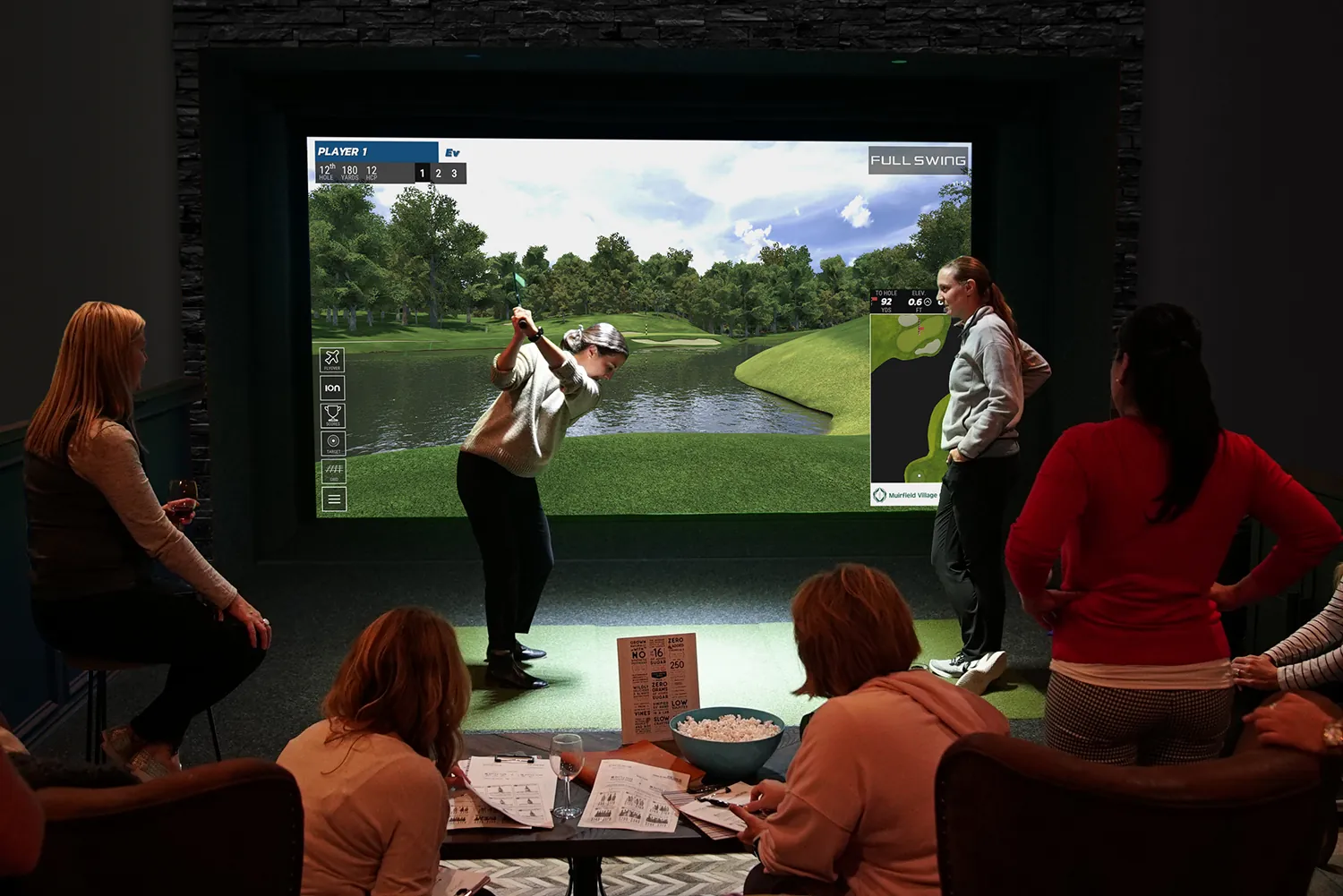 Group of friends enjoying an indoor golf simulator, one taking a swing while the others watch from the lounge seating